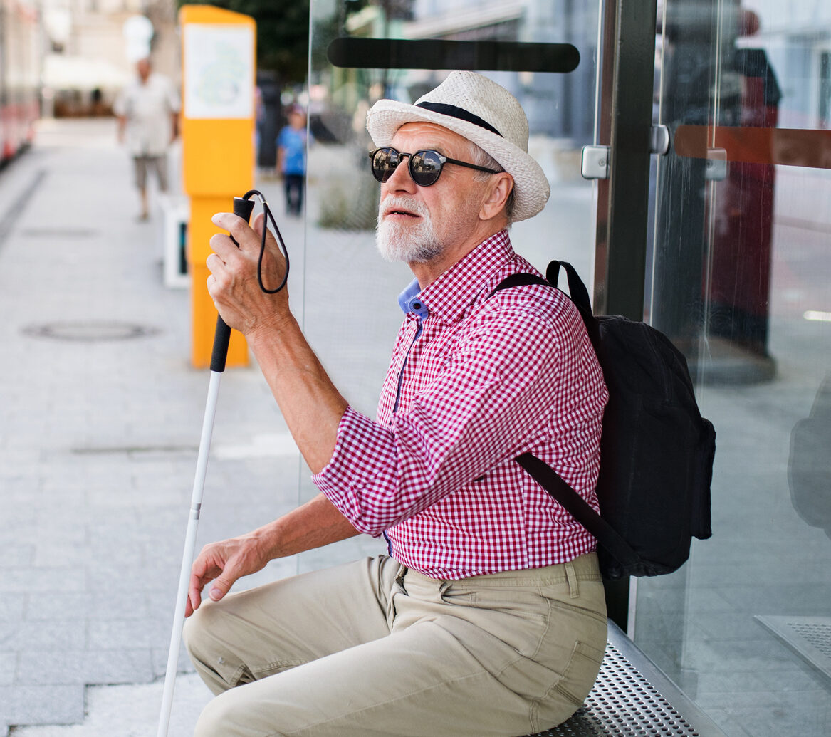 Senior blind man with white cane waiting for public transport in city.
