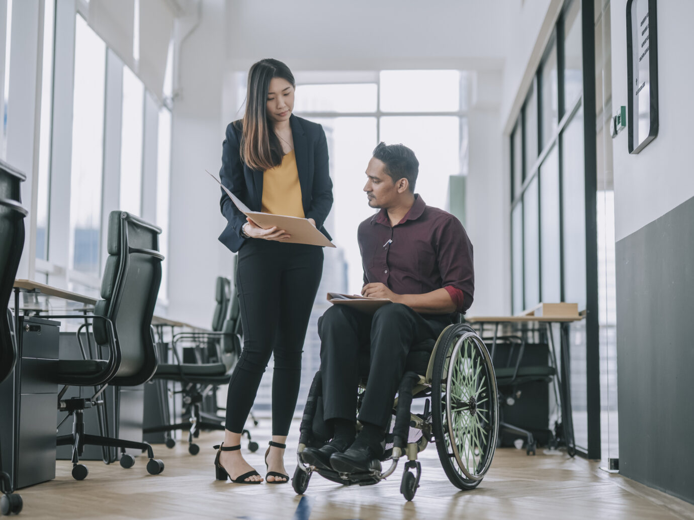 Indian white collar male worker in wheelchair having serious discussion conversation with his female asain chinese colleague coworking at walkway corridor